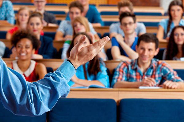 Large group of students sitting in the lecture hall at university and listening to their teacher. Focus on the professor's hand.